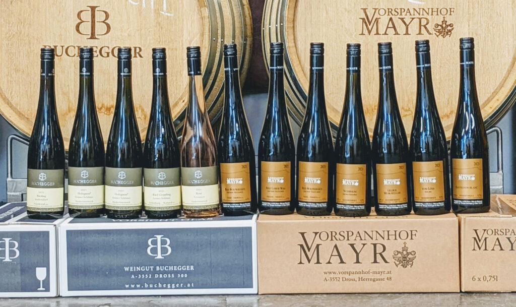 Zwölf Flaschen Wein, dahinter zwei Holzfässer
